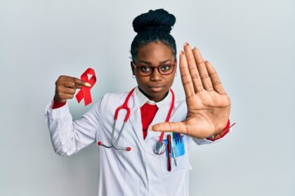 Young african american woman wearing doctor uniform holding support hiv red ribbon with open hand doing stop sign with serious and confident expression, defense gesture